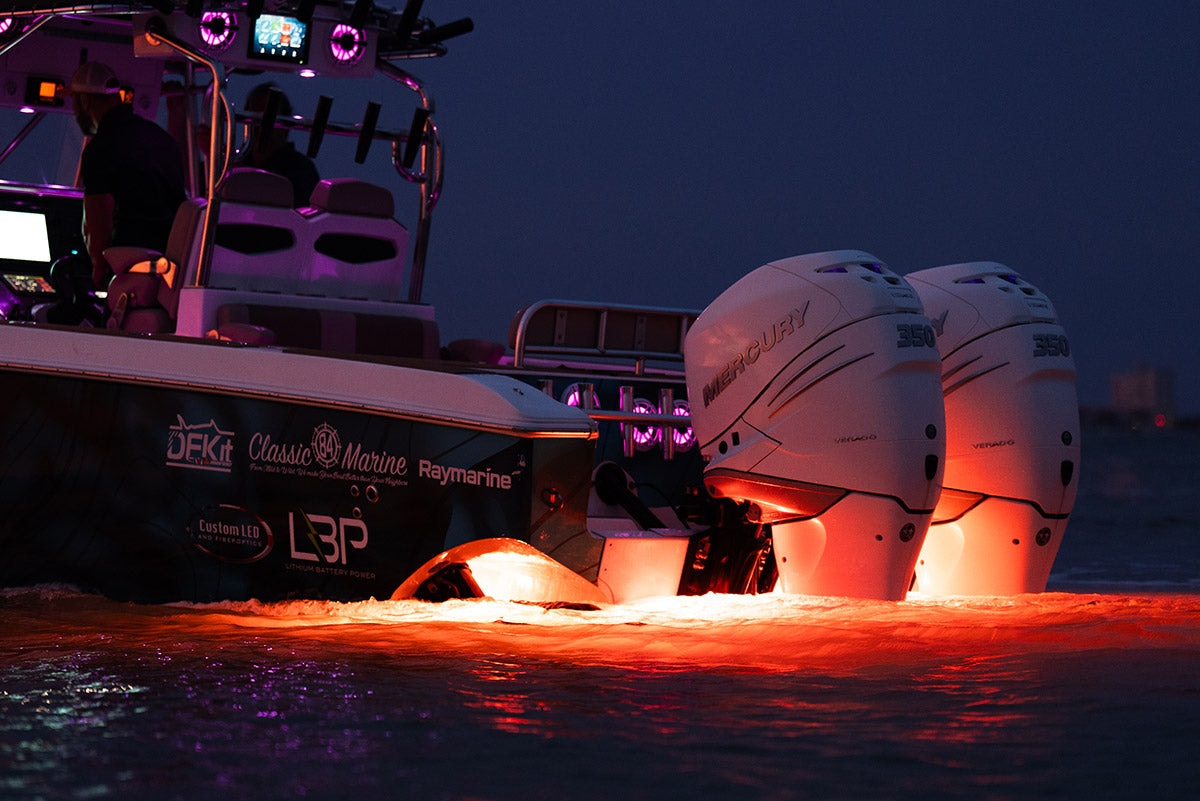A dark speedboat with two white outboard motors at night, illuminated by bright orange underglow reflecting on the water. The boat features visible branding including DPK Marine, Classic Marine, and Raymarine.
