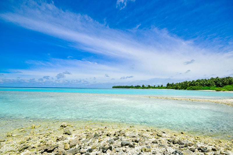 Clear turquoise waters and rocky shore with a tropical island in the distance under a blue sky.