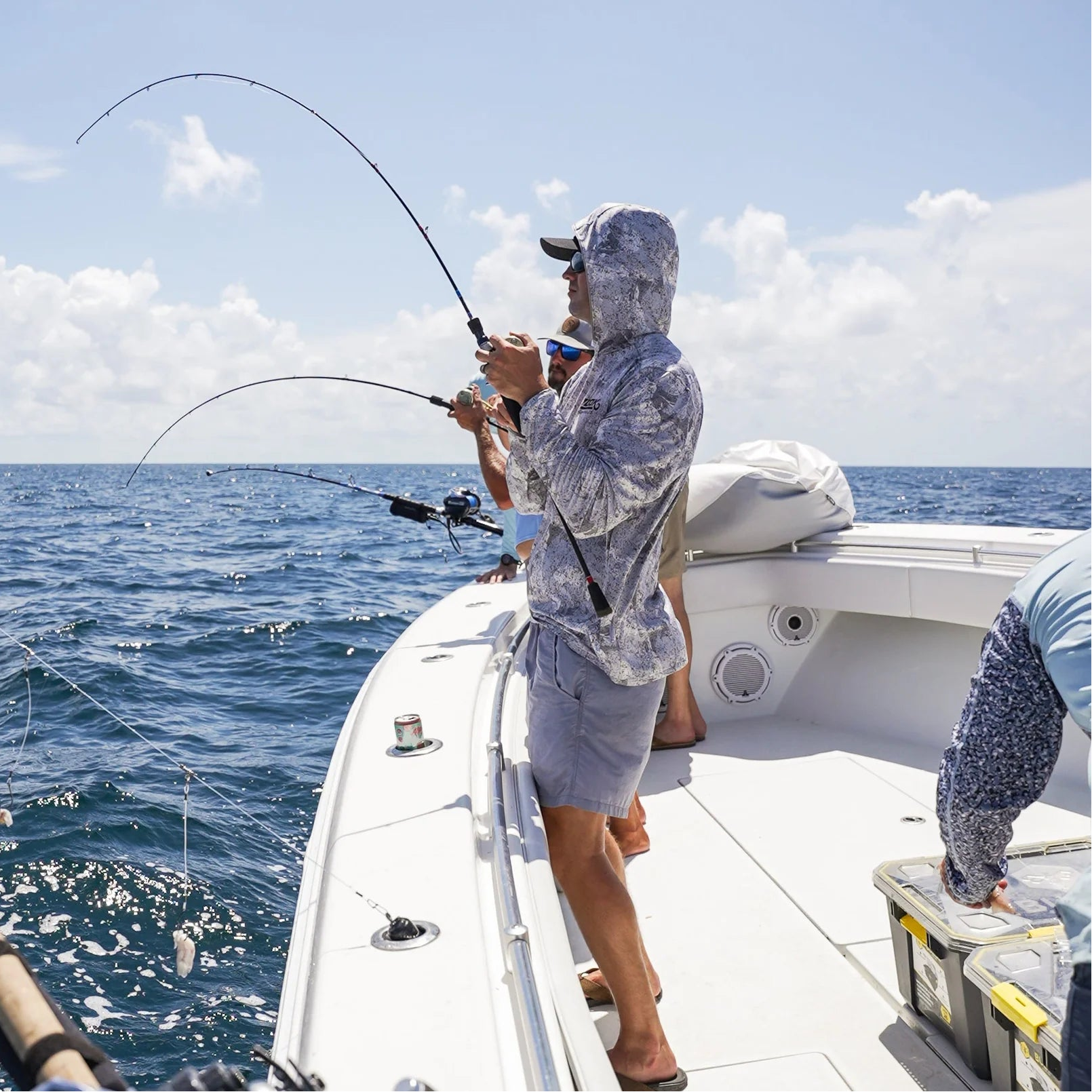 A person stands on a white fishing boat holding a bent fishing rod, with blue ocean water and a partly cloudy sky in the background. Another person is partially visible at the right edge.